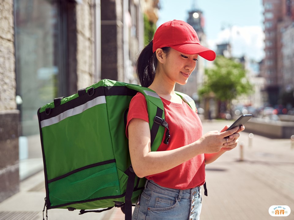 除了網路行銷策略之外，實體的活動也不可忽視。一個亞洲女孩，揹著餐點外送包包，看著手機，確認外送地址。右下角有喜得的LOGO